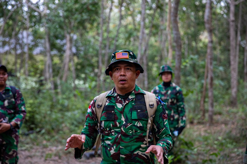 A military personnel wearing Indonesian Army (TNI) camouflage uniform and a bucket hat with a US flag patch is speaking or briefing in a forested area. He is equipped with a tactical vest and backpack, suggesting a field exercise or joint military operation. Two additional soldiers in matching TNI camouflage uniforms are visible in the background.