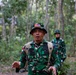 A military personnel wearing Indonesian Army (TNI) camouflage uniform and a bucket hat with a US flag patch is speaking or briefing in a forested area. He is equipped with a tactical vest and backpack, suggesting a field exercise or joint military operation. Two additional soldiers in matching TNI camouflage uniforms are visible in the background.