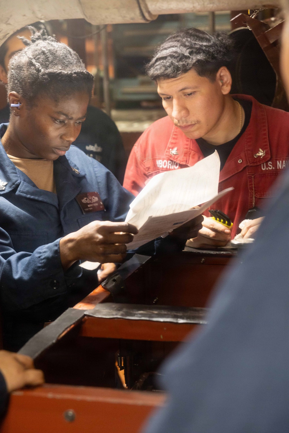 Two U.S. Navy personnel are reviewing a document together in what appears to be a shipboard interior space. The sailor on the left is wearing a blue Navy uniform, while the sailor on the right is wearing a red coverall jacket with 'U.S. NAVY' and a partial name tag reading 'BORIO' visible. They appear to be conducting an inspection or reviewing technical documentation.