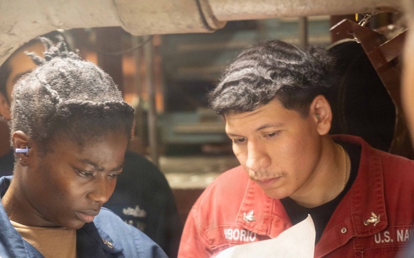 Two U.S. Navy personnel are reviewing a document together in what appears to be a shipboard interior space. The sailor on the left is wearing a blue Navy uniform, while the sailor on the right is wearing a red coverall jacket with 'U.S. NAVY' and a partial name tag reading 'BORIO' visible. They appear to be conducting an inspection or reviewing technical documentation.