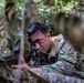 A soldier in multicam/OCP camouflage uniform is positioned in a prone or low-crawl stance concealed within dense vegetation. The individual is aiming down the sights of a rifle, suggesting a tactical concealment or reconnaissance exercise. A unit patch is visible on the right shoulder of the uniform.