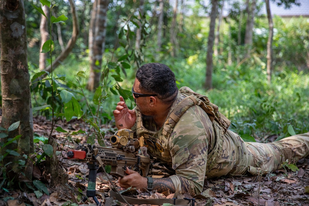 A soldier in full camouflage gear is lying prone on a forest floor, positioned behind a rifle with an optical scope. He appears to be in a tactical observation or overwatch position, with one hand raised in what appears to be a hand signal gesture. The setting is a dense tropical or subtropical woodland environment.