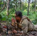 A soldier in full camouflage gear is lying prone on a forest floor, positioned behind a rifle with an optical scope. He appears to be in a tactical observation or overwatch position, with one hand raised in what appears to be a hand signal gesture. The setting is a dense tropical or subtropical woodland environment.