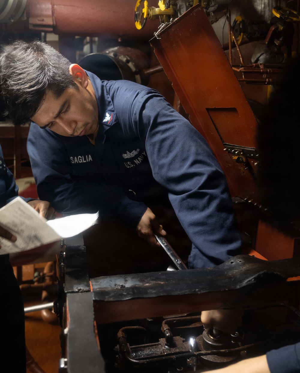 A U.S. Navy sailor with the name tag 'SAGLIA' is performing maintenance work in what appears to be an engine room or mechanical space aboard a naval vessel. The sailor is wearing a navy blue coverall uniform with a U.S. Navy insignia and is using a wrench on machinery. A second person is partially visible holding a document, likely a maintenance procedure or checklist.