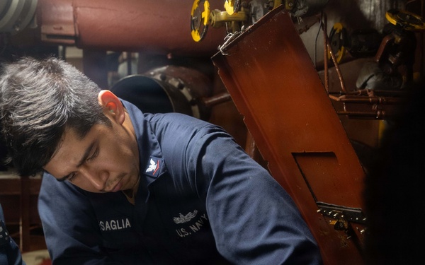 A U.S. Navy sailor with the name tag 'SAGLIA' is performing maintenance work in what appears to be an engine room or mechanical space aboard a naval vessel. The sailor is wearing a navy blue coverall uniform with a U.S. Navy insignia and is using a wrench on machinery. A second person is partially visible holding a document, likely a maintenance procedure or checklist.