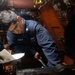 A U.S. Navy sailor with the name tag 'SAGLIA' is performing maintenance work in what appears to be an engine room or mechanical space aboard a naval vessel. The sailor is wearing a navy blue coverall uniform with a U.S. Navy insignia and is using a wrench on machinery. A second person is partially visible holding a document, likely a maintenance procedure or checklist.