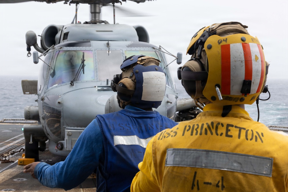 Two naval personnel aboard the USS Princeton are observing a military helicopter on the flight deck of a naval vessel at sea. The crew members are wearing standard flight deck safety gear including cranial helmets with hearing protection and color-coded jerseys indicating their roles. The scene depicts routine flight deck operations with a gray military helicopter positioned in front of them.