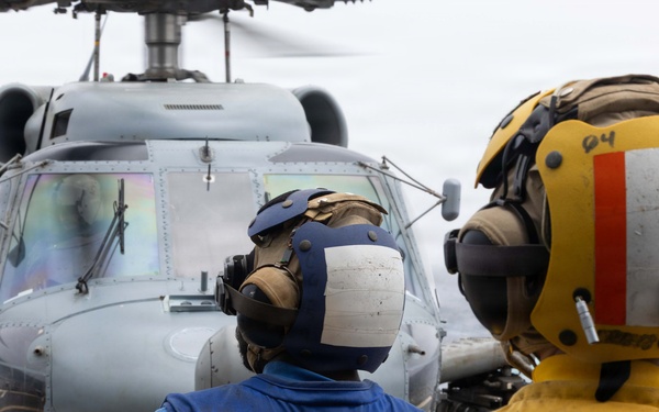 Two naval personnel aboard the USS Princeton are observing a military helicopter on the flight deck of a naval vessel at sea. The crew members are wearing standard flight deck safety gear including cranial helmets with hearing protection and color-coded jerseys indicating their roles. The scene depicts routine flight deck operations with a gray military helicopter positioned in front of them.
