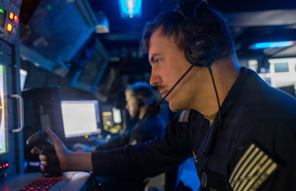 A military or naval operator is seated at a control station in a dimly lit operations room, wearing an EPOS headset with a boom microphone. He is focused on a monitor while operating controls, including what appears to be a joystick. A second operator is visible in the background at an adjacent workstation.