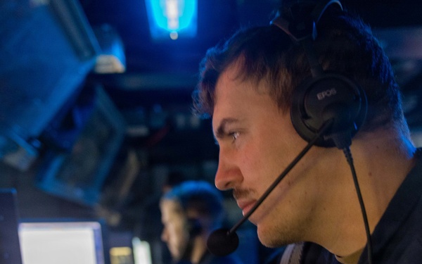 A military or naval operator is seated at a control station in a dimly lit operations room, wearing an EPOS headset with a boom microphone. He is focused on a monitor while operating controls, including what appears to be a joystick. A second operator is visible in the background at an adjacent workstation.