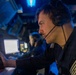 A military or naval operator is seated at a control station in a dimly lit operations room, wearing an EPOS headset with a boom microphone. He is focused on a monitor while operating controls, including what appears to be a joystick. A second operator is visible in the background at an adjacent workstation.