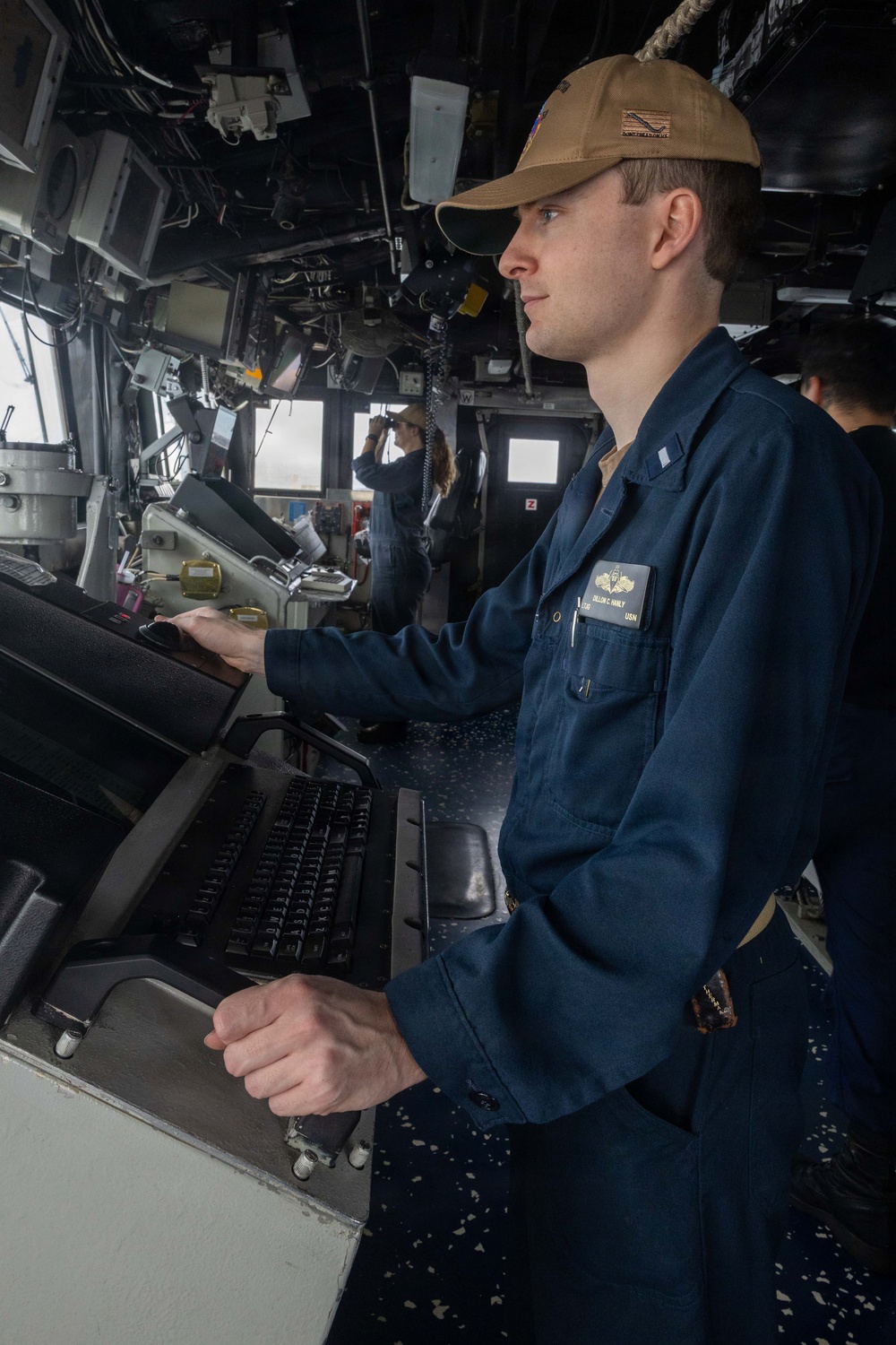A U.S. Navy sailor in blue coveralls (NWU Type III working uniform) stands at a navigation or combat systems console aboard a naval vessel's bridge. The sailor's name tag reads 'DILLON C. FAMILY' with 'USN' below, and they wear a tan/coyote brown ball cap with a single silver bar insignia visible on the collar indicating the rank of Ensign (O-1). In the background, a second crew member is using binoculars while looking out the bridge windows.