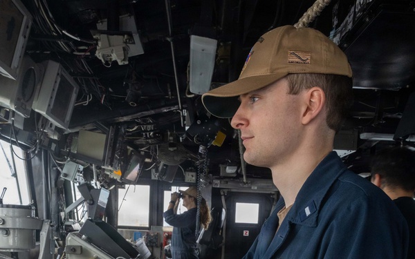 A U.S. Navy sailor in blue coveralls (NWU Type III working uniform) stands at a navigation or combat systems console aboard a naval vessel's bridge. The sailor's name tag reads 'DILLON C. FAMILY' with 'USN' below, and they wear a tan/coyote brown ball cap with a single silver bar insignia visible on the collar indicating the rank of Ensign (O-1). In the background, a second crew member is using binoculars while looking out the bridge windows.