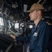 A U.S. Navy sailor in blue coveralls (NWU Type III working uniform) stands at a navigation or combat systems console aboard a naval vessel's bridge. The sailor's name tag reads 'DILLON C. FAMILY' with 'USN' below, and they wear a tan/coyote brown ball cap with a single silver bar insignia visible on the collar indicating the rank of Ensign (O-1). In the background, a second crew member is using binoculars while looking out the bridge windows.