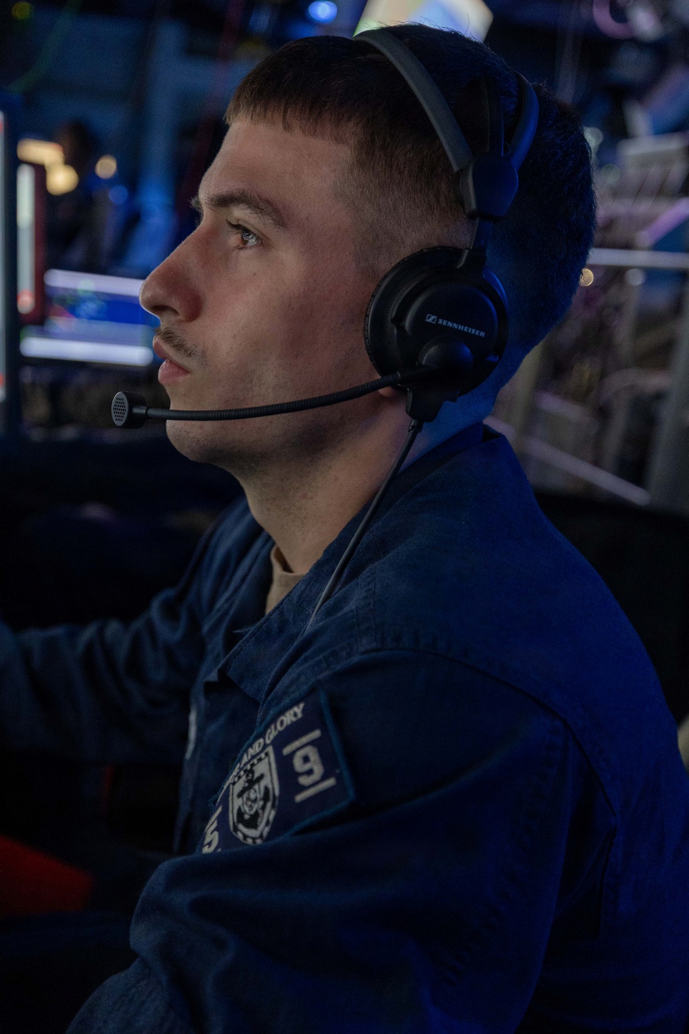 A young man is seated in what appears to be a dimly lit operations or command center environment, wearing a Sennheiser headset with a boom microphone. He is dressed in a dark blue uniform with a patch reading 'AND GLORY' and the number '9' visible on the sleeve. He appears focused and attentive, looking forward in a professional setting.