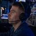 A young man is seated in what appears to be a dimly lit operations or command center environment, wearing a Sennheiser headset with a boom microphone. He is dressed in a dark blue uniform with a patch reading 'AND GLORY' and the number '9' visible on the sleeve. He appears focused and attentive, looking forward in a professional setting.