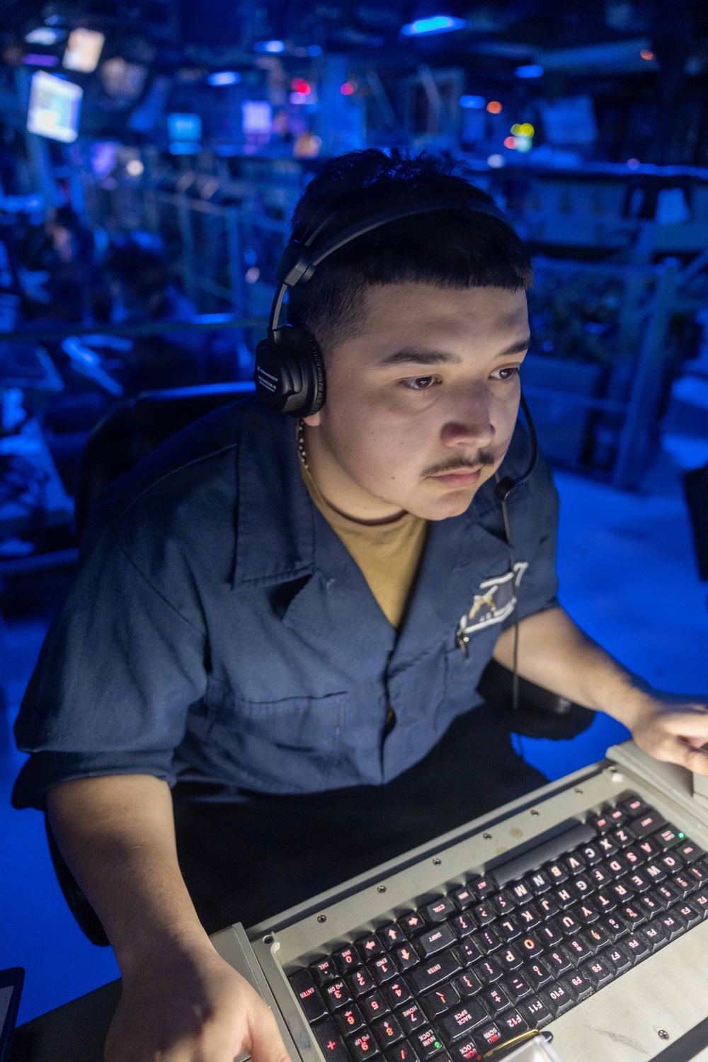A young male sailor in a blue Navy working uniform is operating a ruggedized military keyboard console in what appears to be a combat information center (CIC) or operations room aboard a naval vessel. He is wearing over-ear headphones with a boom microphone and is focused intently on the equipment in front of him. The environment is illuminated in blue tactical lighting, typical of shipboard operations spaces.