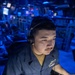 A young male sailor in a blue Navy working uniform is operating a ruggedized military keyboard console in what appears to be a combat information center (CIC) or operations room aboard a naval vessel. He is wearing over-ear headphones with a boom microphone and is focused intently on the equipment in front of him. The environment is illuminated in blue tactical lighting, typical of shipboard operations spaces.