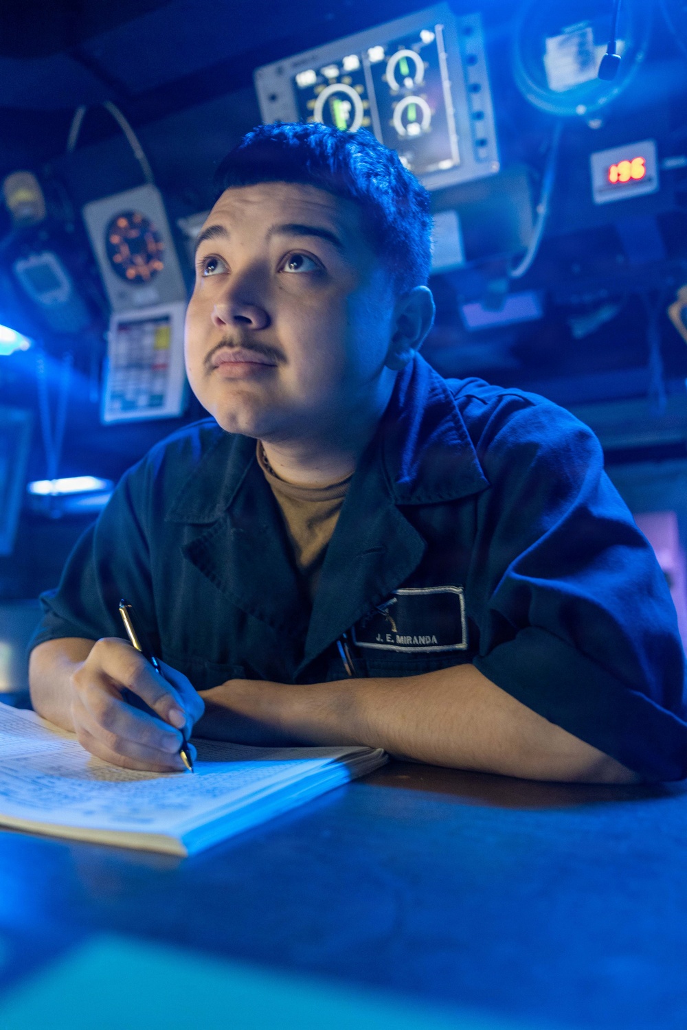 A U.S. Navy sailor identified by name tag as 'J. E. Miranda' is writing in a logbook or notebook while looking upward, likely on the bridge or combat information center of a naval vessel. The environment is illuminated predominantly in blue light, characteristic of a ship's darkened bridge or operations room. Multiple electronic instruments, displays, and control panels are visible in the background.