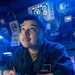 A U.S. Navy sailor identified by name tag as 'J. E. Miranda' is writing in a logbook or notebook while looking upward, likely on the bridge or combat information center of a naval vessel. The environment is illuminated predominantly in blue light, characteristic of a ship's darkened bridge or operations room. Multiple electronic instruments, displays, and control panels are visible in the background.