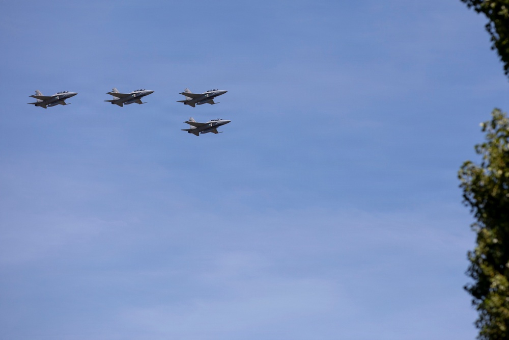 Four military jet aircraft are flying in a loose diamond or finger-four formation against a clear blue sky. The jets appear to be twin-engine fighter aircraft, likely participating in a flyover or air show. A tree canopy is partially visible in the lower right corner of the frame.
