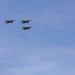 Four military jet aircraft are flying in a loose diamond or finger-four formation against a clear blue sky. The jets appear to be twin-engine fighter aircraft, likely participating in a flyover or air show. A tree canopy is partially visible in the lower right corner of the frame.