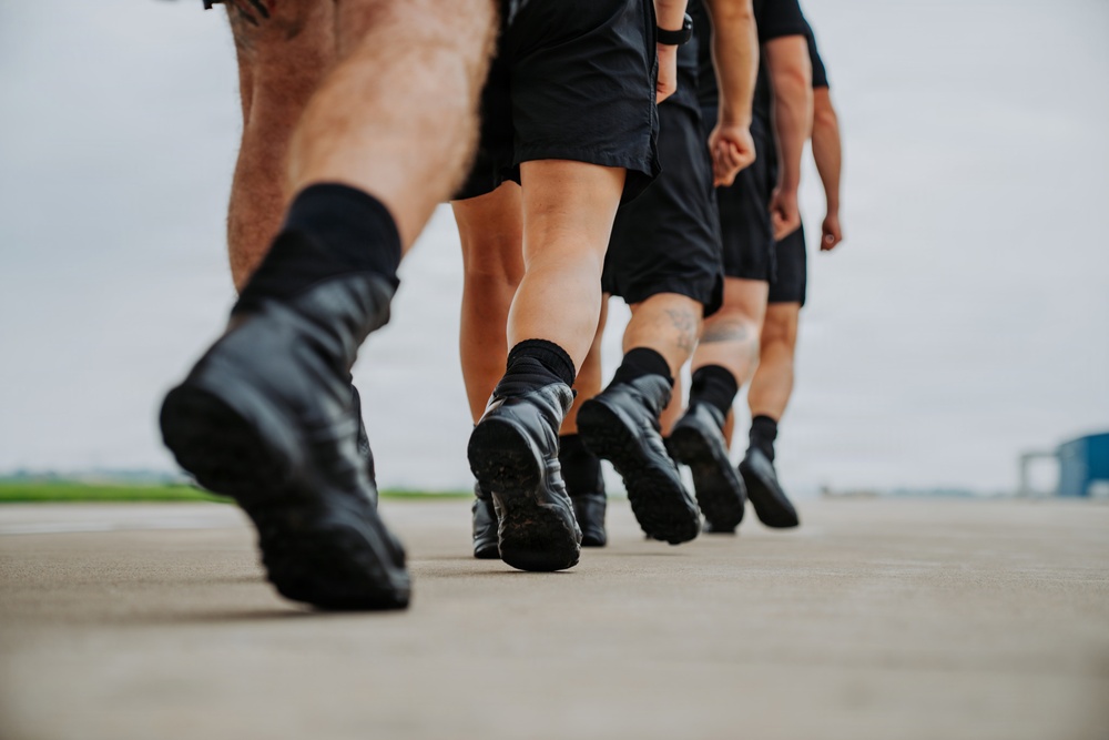 A low-angle close-up photograph showing the lower bodies of multiple individuals marching in formation on a paved surface. All individuals are wearing matching black shorts, black socks, and black boots or athletic shoes, suggesting a military, law enforcement, or paramilitary training context. The depth-of-field effect emphasizes the lead person's boots while the others recede into a blurred background.