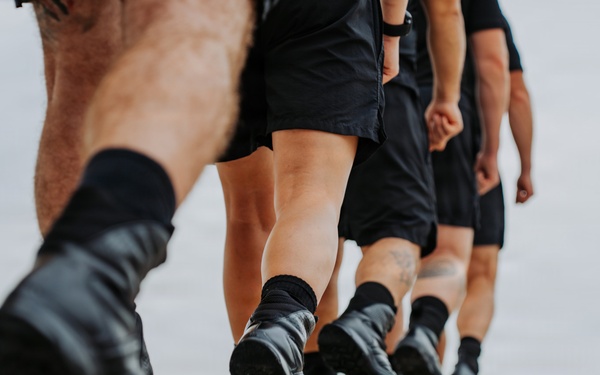 A low-angle close-up photograph showing the lower bodies of multiple individuals marching in formation on a paved surface. All individuals are wearing matching black shorts, black socks, and black boots or athletic shoes, suggesting a military, law enforcement, or paramilitary training context. The depth-of-field effect emphasizes the lead person's boots while the others recede into a blurred background.