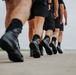 A low-angle close-up photograph showing the lower bodies of multiple individuals marching in formation on a paved surface. All individuals are wearing matching black shorts, black socks, and black boots or athletic shoes, suggesting a military, law enforcement, or paramilitary training context. The depth-of-field effect emphasizes the lead person's boots while the others recede into a blurred background.