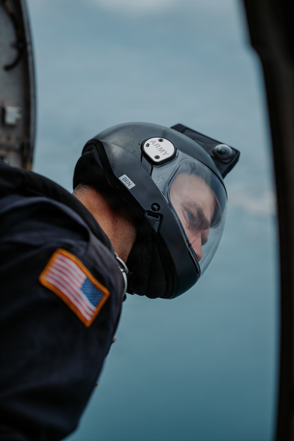 A U.S. Army aviator wearing a black flight helmet with a clear visor is photographed from behind and above, appearing to look downward out of an aircraft. The helmet bears labels reading 'ARMY' and 'BEN', and the subject's flight suit displays a reverse U.S. flag patch with gold border on the left sleeve. The background shows a pale blue sky or water surface, suggesting the aircraft is airborne.