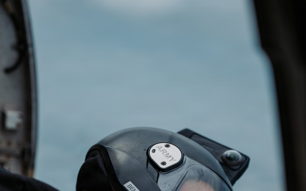 A U.S. Army aviator wearing a black flight helmet with a clear visor is photographed from behind and above, appearing to look downward out of an aircraft. The helmet bears labels reading 'ARMY' and 'BEN', and the subject's flight suit displays a reverse U.S. flag patch with gold border on the left sleeve. The background shows a pale blue sky or water surface, suggesting the aircraft is airborne.