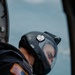 A U.S. Army aviator wearing a black flight helmet with a clear visor is photographed from behind and above, appearing to look downward out of an aircraft. The helmet bears labels reading 'ARMY' and 'BEN', and the subject's flight suit displays a reverse U.S. flag patch with gold border on the left sleeve. The background shows a pale blue sky or water surface, suggesting the aircraft is airborne.