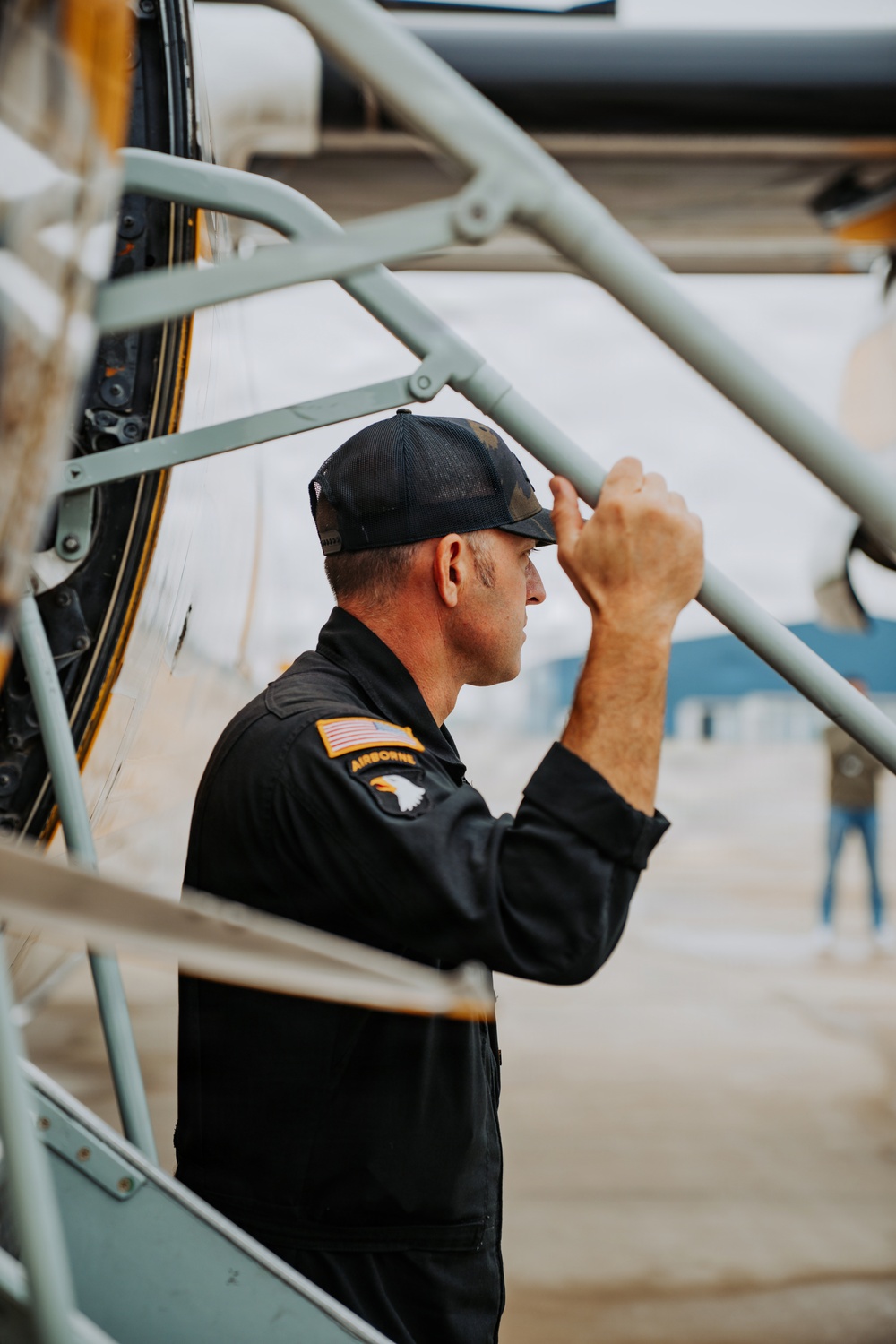 A man wearing a black flight suit with a US flag patch and '101st Airborne' insignia is inspecting or examining an aircraft up close. He is leaning in toward the fuselage of what appears to be a vintage or military aircraft, gripping a structural frame member. The setting appears to be an airfield or air show environment.
