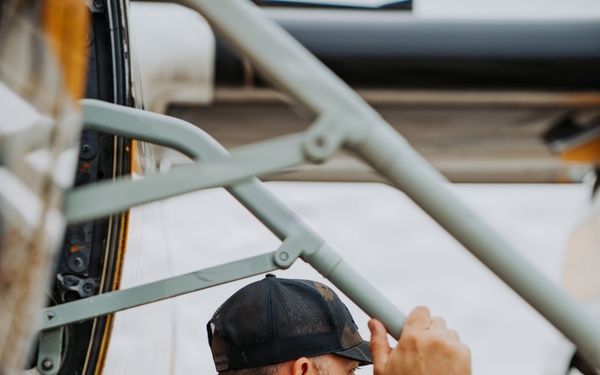 A man wearing a black flight suit with a US flag patch and '101st Airborne' insignia is inspecting or examining an aircraft up close. He is leaning in toward the fuselage of what appears to be a vintage or military aircraft, gripping a structural frame member. The setting appears to be an airfield or air show environment.