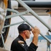 A man wearing a black flight suit with a US flag patch and '101st Airborne' insignia is inspecting or examining an aircraft up close. He is leaning in toward the fuselage of what appears to be a vintage or military aircraft, gripping a structural frame member. The setting appears to be an airfield or air show environment.
