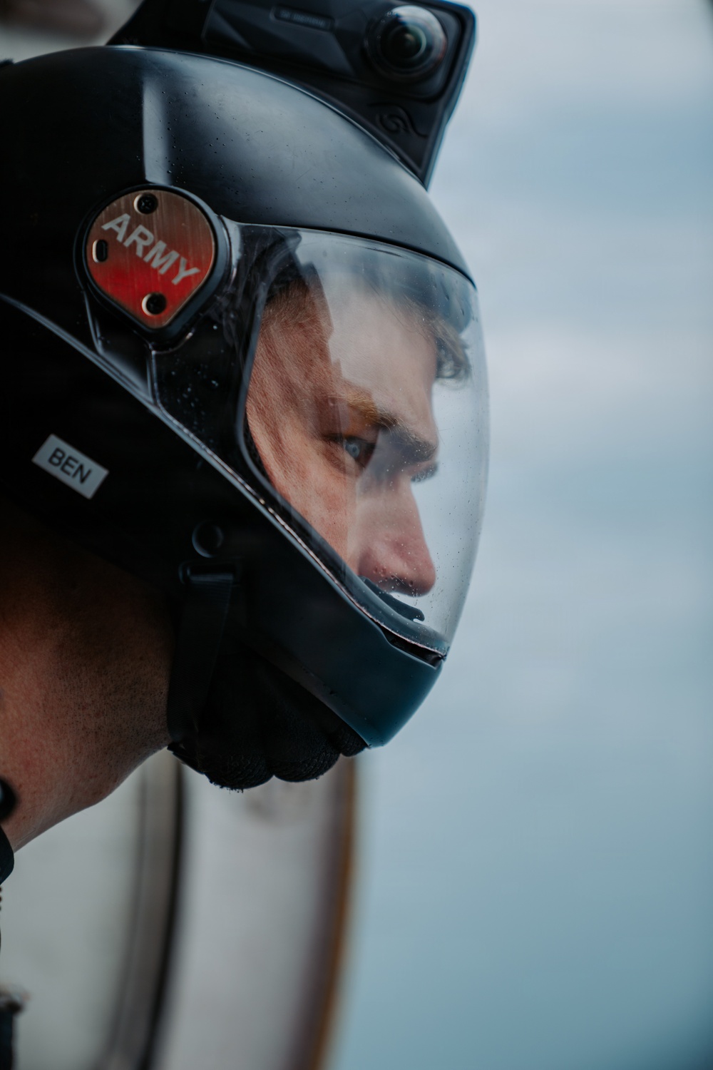A close-up portrait of a person wearing a black full-face helmet with a clear visor. The helmet features a red badge labeled 'ARMY' on the side and a white name tag reading 'BEN', along with a mounted camera on top. The subject's face is partially visible through the visor against a blurred, overcast sky background.