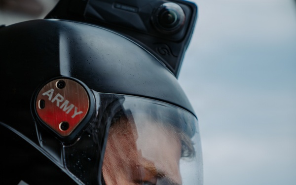 A close-up portrait of a person wearing a black full-face helmet with a clear visor. The helmet features a red badge labeled 'ARMY' on the side and a white name tag reading 'BEN', along with a mounted camera on top. The subject's face is partially visible through the visor against a blurred, overcast sky background.