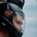 A close-up portrait of a person wearing a black full-face helmet with a clear visor. The helmet features a red badge labeled 'ARMY' on the side and a white name tag reading 'BEN', along with a mounted camera on top. The subject's face is partially visible through the visor against a blurred, overcast sky background.