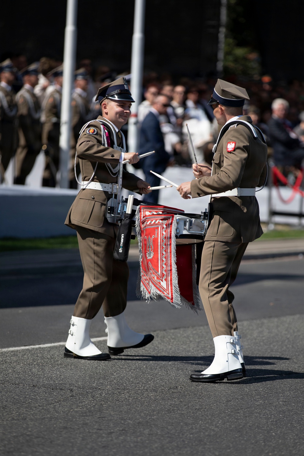 Two Polish military band drummers are performing during a formal parade or ceremonial event. They are facing each other while playing snare drums adorned with red decorative aprons featuring embroidered insignia. A crowd of spectators and additional uniformed soldiers are visible in the background.