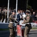 Two Polish military band drummers are performing during a formal parade or ceremonial event. They are facing each other while playing snare drums adorned with red decorative aprons featuring embroidered insignia. A crowd of spectators and additional uniformed soldiers are visible in the background.