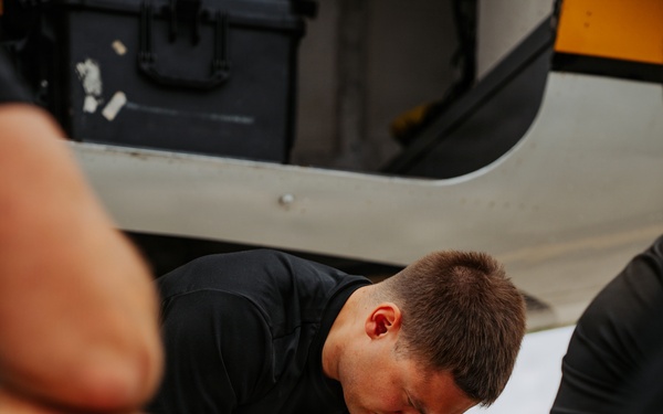 A young man wearing a black uniform with a military-style patch is kneeling on a tarmac, using a yellow cordless power drill on components near what appears to be an aircraft. He is surrounded by other individuals and various tools and materials including zip ties and mechanical components. The setting appears to be an airfield or flight line.