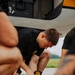 A young man wearing a black uniform with a military-style patch is kneeling on a tarmac, using a yellow cordless power drill on components near what appears to be an aircraft. He is surrounded by other individuals and various tools and materials including zip ties and mechanical components. The setting appears to be an airfield or flight line.