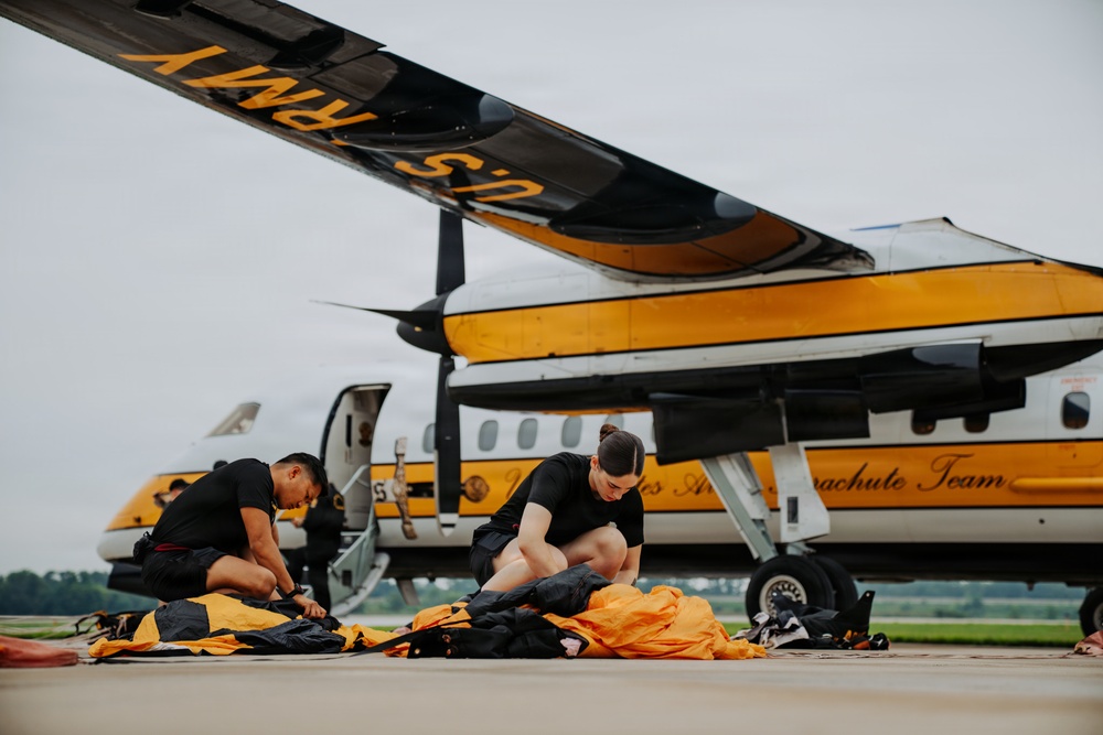 Two individuals dressed in black uniforms are packing or preparing parachutes on an airport tarmac in front of a U.S. Army Golden Knights parachute team aircraft. The plane is distinctively painted in yellow and white with 'U.S. Army' and 'Parachute Team' text visible on the fuselage. The scene appears to be pre-jump preparation at an airshow or military demonstration event.