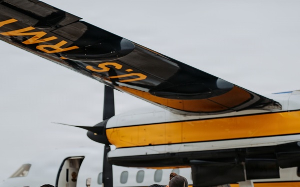 Two individuals dressed in black uniforms are packing or preparing parachutes on an airport tarmac in front of a U.S. Army Golden Knights parachute team aircraft. The plane is distinctively painted in yellow and white with 'U.S. Army' and 'Parachute Team' text visible on the fuselage. The scene appears to be pre-jump preparation at an airshow or military demonstration event.