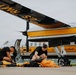 Two individuals dressed in black uniforms are packing or preparing parachutes on an airport tarmac in front of a U.S. Army Golden Knights parachute team aircraft. The plane is distinctively painted in yellow and white with 'U.S. Army' and 'Parachute Team' text visible on the fuselage. The scene appears to be pre-jump preparation at an airshow or military demonstration event.
