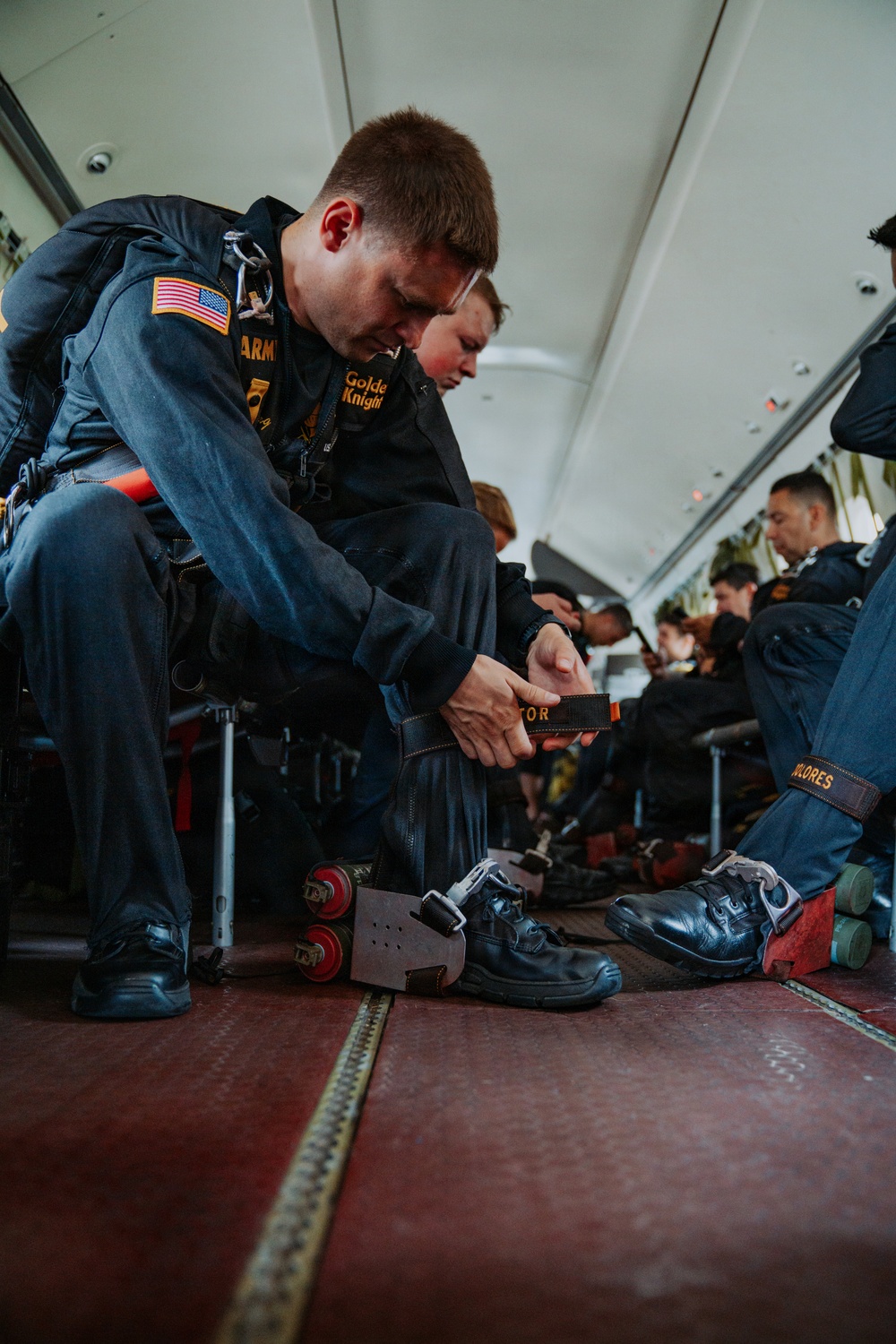 Members of the U.S. Army Golden Knights parachute team are seated inside a military aircraft, preparing their equipment before a jump. The foreground soldier is adjusting a strap or harness component while specialized jump equipment, including what appear to be smoke canisters and a board-like device, are visible on the floor. Multiple team members in matching dark blue jumpsuits with gold insignia are visible in the background doing similar pre-jump preparations.