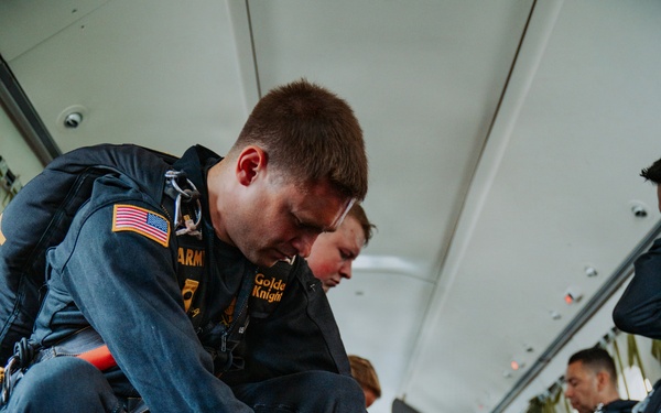 Members of the U.S. Army Golden Knights parachute team are seated inside a military aircraft, preparing their equipment before a jump. The foreground soldier is adjusting a strap or harness component while specialized jump equipment, including what appear to be smoke canisters and a board-like device, are visible on the floor. Multiple team members in matching dark blue jumpsuits with gold insignia are visible in the background doing similar pre-jump preparations.