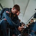 Members of the U.S. Army Golden Knights parachute team are seated inside a military aircraft, preparing their equipment before a jump. The foreground soldier is adjusting a strap or harness component while specialized jump equipment, including what appear to be smoke canisters and a board-like device, are visible on the floor. Multiple team members in matching dark blue jumpsuits with gold insignia are visible in the background doing similar pre-jump preparations.