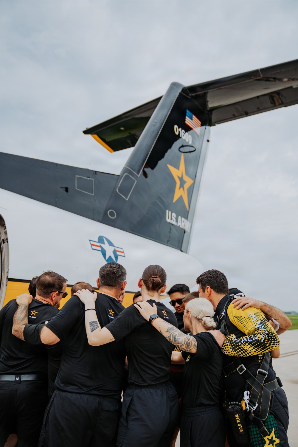A group of U.S. Army personnel in black uniforms are gathered in a team huddle on an airfield tarmac beside a U.S. Army aircraft. The tail of the aircraft displays the serial number '01609,' a gold star emblem, 'U.S. ARMY' text, and an American flag decal. At least one member is wearing a parachute harness and a yellow helmet marked 'GOLD TEAM,' indicating this is likely the U.S. Army Golden Knights parachute demonstration team.