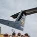 A group of U.S. Army personnel in black uniforms are gathered in a team huddle on an airfield tarmac beside a U.S. Army aircraft. The tail of the aircraft displays the serial number '01609,' a gold star emblem, 'U.S. ARMY' text, and an American flag decal. At least one member is wearing a parachute harness and a yellow helmet marked 'GOLD TEAM,' indicating this is likely the U.S. Army Golden Knights parachute demonstration team.